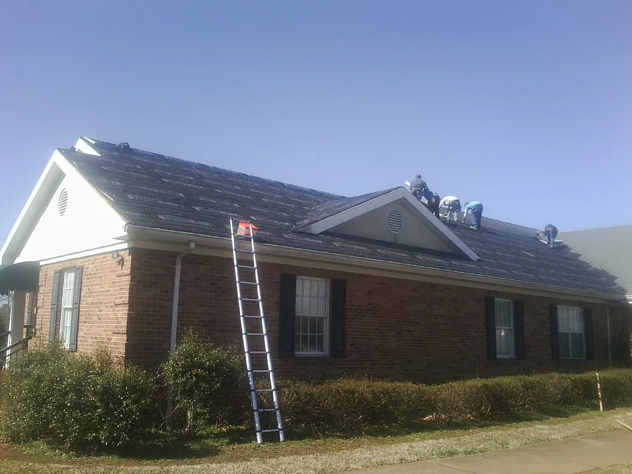 Crew tearing shingles off a brick ranch roof with a ladder set — Metal Roof Repair work by our crew in Evansdale