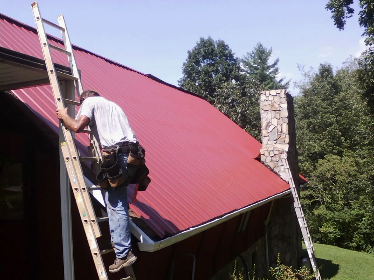 Roofer on a ladder beside a red metal roof and stone chimney — Soffit & Fascia Repair by Roofing Brothers in Evansdale